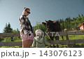 young woman strokes head brown cow sleeve, little girl nearby tries get closer look through fence afraid. Later, another cow approaches quiet, lazy summer day country village, encounter farm animals. 143076123