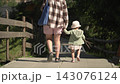 A little girl pink dress carries bundle sticks, she takes her mother's hand look concentration. Shot from behind, family walks along a paved path leading down into the forest on a warm, sunny day. 143076124