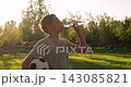 Young boy drinks water and holds soccer ball in sunny park. Child athlete takes refreshing break after football practice. Golden sunlight creates healthy outdoor mood. Hydration supports fitness. 143085821