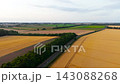 Empty country road winding between golden wheat fields and green woodland at sunset. Aerial drone view over agricultural countryside with harvested farmland. Peaceful rural landscape under soft 143088268