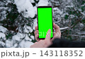 Young man's hand holding smartphone with green screen against snowy forest background. Vertical mockup of mobile phone. Man using phone with blank green display background. Smartphone with chroma key 143118352