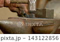 Macro closeup shot captures medieval blacksmith striking hot metal with heavy hammer on cast iron anvil in open-air workshop at historical craft fair. 143122856