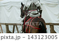 Medium rear view shot shows medieval knight in steel plate armor sitting on brown warhorse with decorative red leather harness by wooden tournament barrier near white camp tents... 143123005