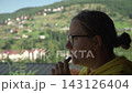 woman yellow hoodie glasses smokes electronic cigarette, exhaling vapor while enjoying view mountains forest warm, cloudless morning. numerous private vacation homes visible mountainside background. 143126404