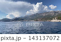 Waterfront resort town rests beneath forested mountain cliffs along blue coastline. Apartment buildings face shore, where small boats sit near beach in scenic alpine setting with forested slopes 143137072