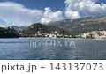 Waterfront resort town rests beneath forested mountain cliffs along blue coastline. Apartment buildings face shore, where small boats sit near beach in scenic alpine setting with forested slopes 143137073