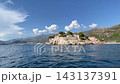 Anchored boats rest on blue harbor water beside historic stone buildings Sveti Stefan with terracotta roofs. Coastal settlement stands along rocky shoreline, while gentle ripples create serene 143137391
