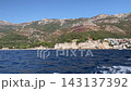 Anchored boats rest on blue harbor water beside historic stone buildings Sveti Stefan with terracotta roofs. Coastal settlement stands along rocky shoreline, while gentle ripples create serene 143137392