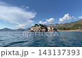 Stone village Sveti Stefan stands above calm Adriatic water, with pale homes and red tile roofs lining rugged rock. Coastal settlement rests beneath distant mountains in bright, peaceful weather 143137393