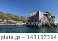 Stone village Sveti Stefan stands above calm Adriatic water, with pale homes and red tile roofs lining rugged rock. Coastal settlement rests beneath distant mountains in bright, peaceful weather 143137394