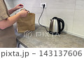 Hand places lidded drink into paper carry bag on kitchen counter. Person prepares kitchen pickup while gripping bag handle and steadying cup inside before carrying packaged beverage for delivery 143137606