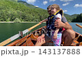 Father helps child hold wooden oars while rowing boat on clear lake. Calm parent keeps young child close in life jacket, sharing sunny family outdoor recreation moment while guiding child to paddle 143137888