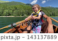 Father embraces young child in life vest while seated aboard wooden boat on calm lake. Protective life jacket and close family moment suit safe boating, with forested hills in sunlight 143137889
