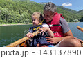 Father holds his young daughter close while seated in wooden boat on sunny lake. Affectionate family pair enjoy boating outdoors, with wooded mountains and rippling water behind them 143137890