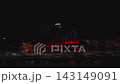 Aerial night view shows the Latvian National Library in Riga with red, blue, and white LED columns, a faint Ferris wheel, car lights, and water reflections during Riga Staro 2025. 143149091
