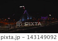 Aerial night view in Riga shows Latvian National Library lit with blue, purple and amber projections, white and red lantern, LED columns, car trails, and riverside promenade 143149092