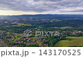 Camera glides over valley suburb, revealing detached homes, apartment blocks, gardens, winding roads, farmland edges and tree belts, with layered hills, haze and heavy clouds above 143170525