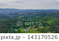 Elevated oblique aerial view showing dense mixed forest, small residential cluster, winding roads, grassy clearings, layered wooded ridges, and meadow pockets across broad rural landscape 143170526