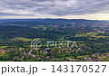Elevated oblique aerial motion scene showing clustered residential neighborhood in foreground transitioning to mixed farmland, pastures, and forest patches in midground, then rising to layered 143170527