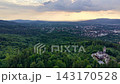 Elevated oblique aerial view showing dense forest canopy in foreground, stone castle estate with tower near lower-right, suburban town across midground, and layered hills with shifting warm light 143170528