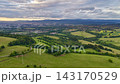 Sweeping rural landscape with layered hills, open pastures, mixed woodland, hedgerows, winding road, patchwork fields, bright crops, and soft horizon glow beneath dramatic cloud cover 143170529