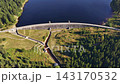 Aerial view of curved dam spanning deep blue lake, surrounded by dense evergreen forest and green slopes, showing dam crest, shoreline curve, and calm water under daylight 143170532