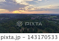 Wide aerial view of sunset glow across patchwork farmland, dense green forest, and small village cluster in valley as clouds part and golden sunbeams stream over rolling hills 143170533