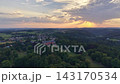 Golden hour aerial panorama of rural valley village with rolling fields, tree forests and distant hills, featuring red roof houses and stone bridge under warm sunset light, long shadows and sky 143170534