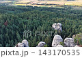 From high altitude, weathered pale rock pillar and jagged outcrop stand over thick pine canopy, opening to patterned fields, farm structures, and long sunset shadows across valley 143170535