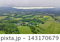 High oblique view over rural valley showing mixed woodland, patchwork fields, yellow flowering crop strips, small homes, winding tracks, rolling hills, low cloud, bright horizon light 143170679