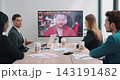 Caucasian executive talks as gestures to online speaker on screen at office call. Diverse business team watches video meeting following discussion attentvely 143191482