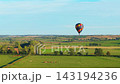 Hot air balloon flying low over green farmland under blue sky. Colorful aircraft above rural fields with trees and grazing cattle. Outdoor aviation scene across agricultural landscape on clear sunny 143194236