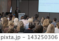 Conference speaker beside slides with listeners in foreground 143250306