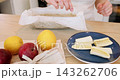 Cake making. Fruit pastry. Female confectioner hand adding banana pieces to dough in baking dish parchment on table with apple orange. 143262706