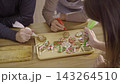 Group of people decorating festive gingerbread biscuits with colorful cream icing piping bags around wooden table during Christmas. Happy caucasian family enjoy baking and decorating gingerbread. 143264510