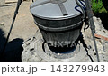 Traditional Cast Iron Cauldron with Lid Hanging on Metal Tripod for Outdoor Cooking Over Fire at Rustic Campsite 143279943