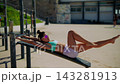 Yoga instructor in pastel sportswear stretches on seaside workout bench 143281913