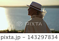 Woman pauses by sunset lakeside, her white hat and coat glowing in golden light. She looks over still water with calm, thoughtful expression as evening reflections shimmer behind her. 143297834