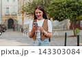 Smiling young woman using smartphone typing texting scrolling social media app on city town street 143303084