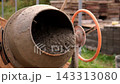 View of concrete mixer drum with wet cement residue, rust and mud on metal surface. Orange hand wheel and support arm beside stacked materials and unfinished masonry outdoors. 143313080