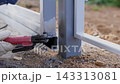 Gloved hand uses welding tool on metal gate joint outdoors, sending bright sparks and smoke from fresh seam beside pale steel posts over gravel ground, with welding, welder, glove, hand detail 143313081