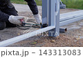 Close-up outdoor repair scene of worker using hand tools to join metal post joint beside coated structure, with bright sparks, gravel texture, and soft daylight 143313083