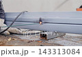 Gloved hands use welding torch on painted metal beam, creating bright white violet flare and dark burn mark at contact point, with welding, flare, spark, torch detail, showing metal, beam, gloves 143313084