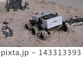 Small wheeled robotic rover moves across pale sandy ground beside dark rock fragments, with white metal chassis, exposed electronics, and textured tires in soft diffuse light, creating rugged 143313393