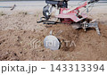 Red and black farm cultivator disturbs dry soil beside paved edge, throwing dust and loose earth across rough foreground, with farm, agriculture, tilling, soil detail, showing dust, cultivator 143313394