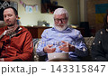 In the living room, an elderly man sits on the couch in a good mood with a bowl of popcorn. He is surrounded by his grandchildren, who are in a bad mood. The grandfather tries to get their attention 143315847