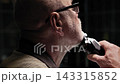 Old man wearing glasses using a trimmer in his hands to shave himself in front of bathroom mirror. The brightly lit head of a man stands in sharp contrast against the dark background of the bathroom 143315852