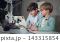 A working 3D printer stands in the foreground of the shot out of focus.Two children in focus sit at a table with a working 3D printer, observing and adjusting its work with laptop. Slow motion. Close 143315854