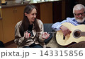 At home, in a brightly lit living room reminiscent of an artistic studio, young woman with long hair and casual clothes sit on the sofa next to their grandfather. She clap along with his guitar 143315856