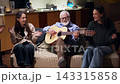 At home, in a brightly lit living room reminiscent of an artistic studio, two young women with long hair and casual clothes sit on the sofa next to their grandfather. They clap along with his guitar 143315858